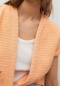 Pfirsichfarbener Strickcardigan mit strukturiertem Muster, offenem Vorderteil und kurzen Ärmeln über einem weißen Tanktop. Weicher, leichter Stoff.