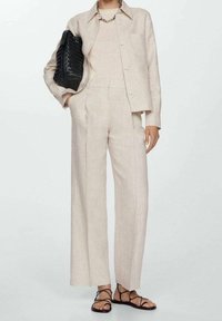 Costume en lin beige avec veste ajustée et pantalon à jambes larges. Le modèle tient un sac à main tissé noir et porte des sandales à lanières. Tissu texturé sur l'ensemble.