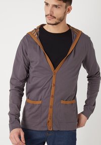 Sudadera gris con un frente abotonado marrón, que cuenta con dos bolsillos frontales y detalles a rayas en la capucha. Está hecha de un tejido suave y ligero.