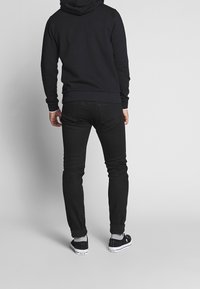 Felpa nera con cappuccio e coulisse, abbinata a jeans slim-fit neri. Il modello indossa sneaker nere con suola bianca e orli risvoltati.