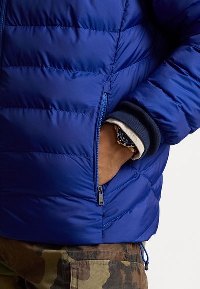 Casaco azul acolchoado com costura horizontal, apresentando um bolso lateral com fecho. Por baixo, é visível um punho azul-escuro e um relógio com mostrador preto.