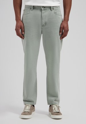 Man draagt lichtgrijze, rechte jeans en taupekleurige suède sneakers met witte veters, staand tegen een effen achtergrond.