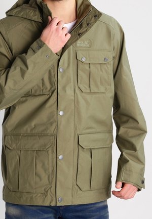 Mann trägt eine olivgrüne Jacke von Jack Wolfskin mit mehreren Fronttaschen und Druckknöpfen und richtet den Kragen.