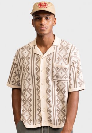 Νεαρός άνδρας που φοράει μπεζ κοντομάνικο πουκάμισο με σχέδια και μπεζ καπέλο με κόκκινο κείμενο και γραφικά.