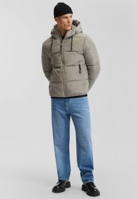 Hellegraue Daunenjacke mit Kapuze, zwei Reißverschluss-Fronttaschen, verstellbaren Zugbändern und leichtem Material. Getragen mit blauen Jeans und schwarzen Schuhen.