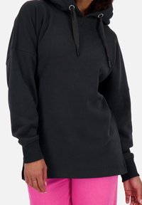 Felpa nera con cappuccio, a taglio ampio, maniche lunghe e spacchi laterali. Presenta dettagli con cordino e polsini a costine. Indossata con pantaloni rosa.