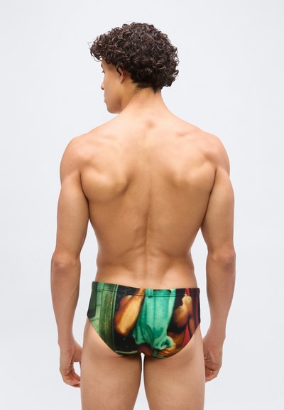 Άνδρας με μπούκλες, εμφανιζόμενος από πίσω, φοράει πολύχρωμο μαγιό με εκτυπωμένη εικόνα ενός ατόμου που κρατάει ένα πράσινο ύφασμα.