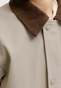 Veste beige avec col en velours côtelé marron côtelé et boutons-pression beiges, portée sur une chemise blanche.
