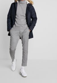 Marinvattensparka med foder av päls, grå turtleneck och grå rutiga byxor. Skorna är vita sneakers med en strukturerad design.