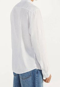 Uomo che indossa una camicia di lino bianca a maniche lunghe e jeans blu, mostrato di spalle e da destra.