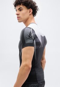 Νεαρός άνδρας με σγουρά μαλλιά, φορώντας εφαρμοστή αθλητική μπλούζα μαύρου και γκρι με μοτίβο, στέκεται πλαγίως μπροστά σε ένα απλό λευκό φόντο.