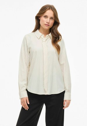 Donna che indossa una blusa a righe color crema con cravatta integrata e pantaloni neri, in piedi di fronte a uno sfondo bianco.
