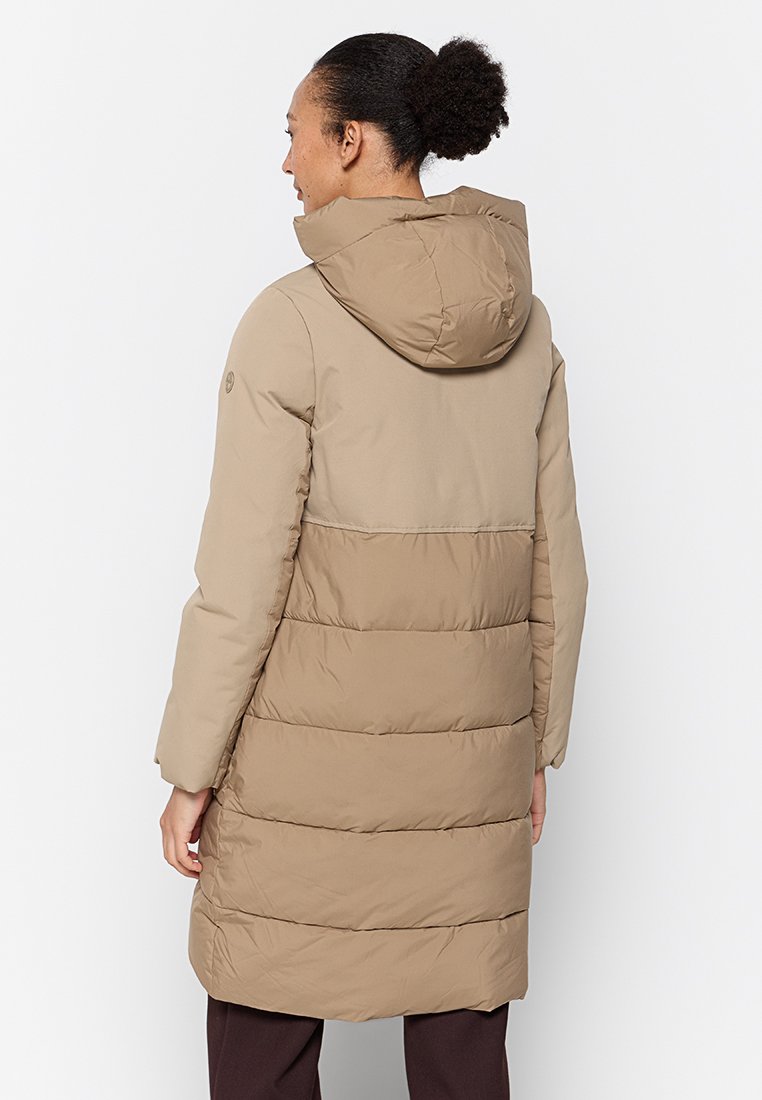 Frau, die einen langen beigefarbenen gepolsterten Mantel mit Kapuze trägt, steht mit dem Rücken zu einem schlichten weißen Hintergrund.