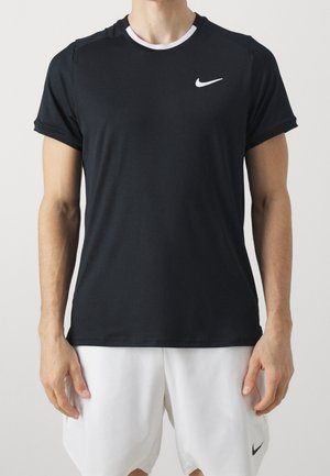 Mandlig torso iført sort Nike kortærmet sportstrøje og hvide Nike shorts, stående mod en ensfarvet lys baggrund.