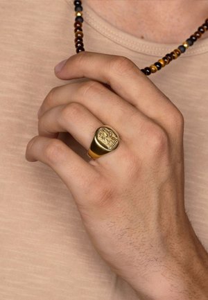 Gold signet ring featuring an embossed design, smooth finish, oval shape, worn on a hand with a beaded dark necklace against a light shirt.