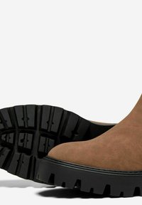 Bottine en suede marron avec un bout arrondi, épaisse semelle en caoutchouc noir et motif de semelle profond pour une meilleure adhérence. Texture lisse, design minimaliste.