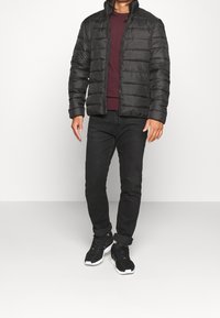 Schwarze Steppjacke mit gesteppter Optik, hohem Kragen und Reißverschluss. Trägt dazu einen weinroten Pullover, schwarze Jeans und schwarze Sneaker.