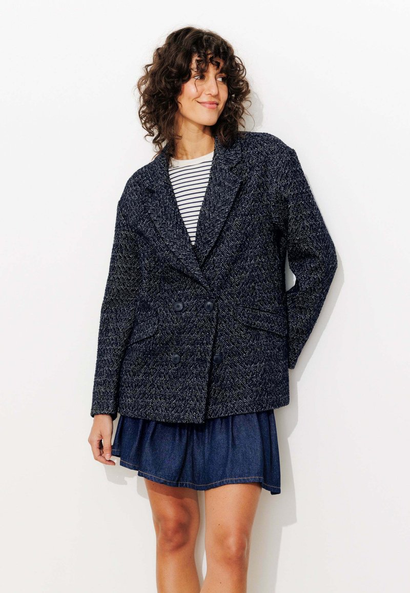 Blazer croisé en tissu texturé bleu marine avec deux poches avant et des boutons noirs, associé à une chemise rayée et une jupe en denim.