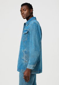 Chaqueta de mezclilla azul con cuello, bolsillos frontales y detalles de puños abotonados. Presenta un corte holgado y una tela texturizada.