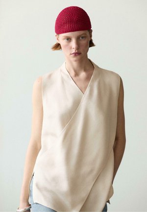 Femme portant un haut beige sans manches cache-cœur et un bonnet rouge au crochet, debout devant un fond clair uni.