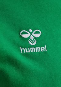 Groene stof met een gladde textuur, met een wit geborduurd logo en de tekst "hummel" aan de onderkant, centraal op de stof gepositioneerd.