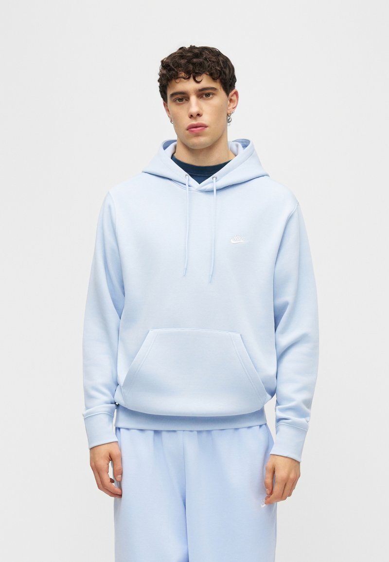 Joven con sudadera con capucha Nike azul claro y pantalones deportivos a juego, posando frente a un fondo gris liso.