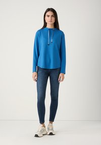 Blauw longsleeve shirt met hoge hals, twee trekkoorden en een relaxte pasvorm, gecombineerd met donker skinny jeans en neutraal gekleurde sneakers.
