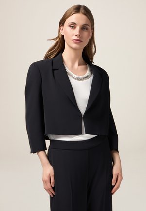 Sacou negru croppat cu o textură netedă, design structurat și mâneci trei sferturi. Asortat cu un top alb ce prezintă o colier argintie.
