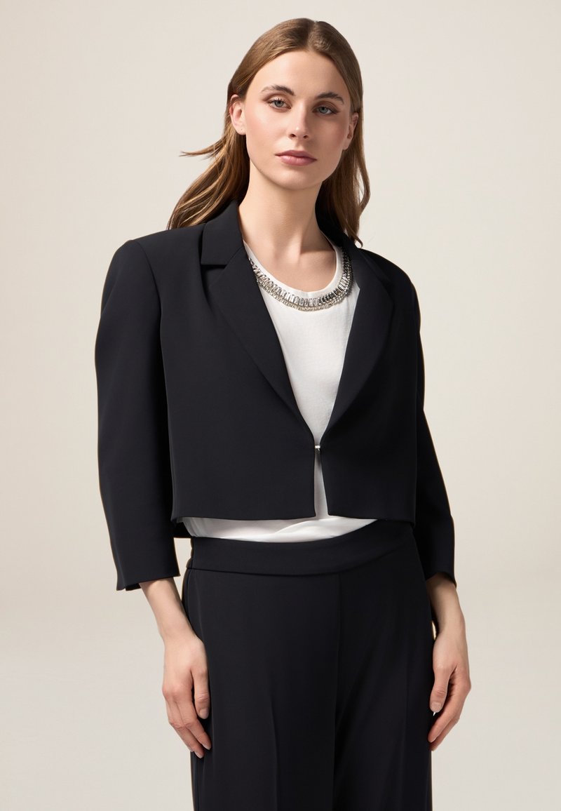 Blazer nero tagliato con una texture liscia, design strutturato e maniche a trequarti. Abbinato a un top bianco con una collana argentata.