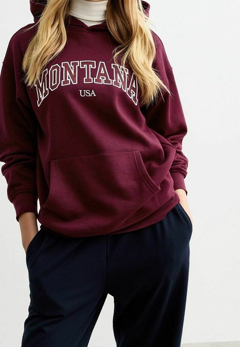 Personne portant un sweat à capuche bordeaux avec le texte « MONTANA USA », superposé à un col roulé blanc, les mains dans les poches d'un pantalon sombre.