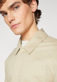 Chaqueta ligera beige con cuello puntiagudo, que presenta una textura suave y botones de presión, superpuesta a un suéter blanco de canalé.