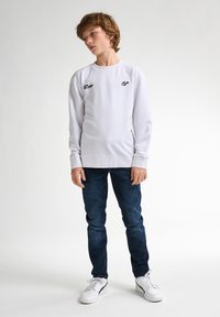 Sudadera blanca de manga larga con estampados de logotipo negro, combinada con vaqueros azul oscuro y zapatillas blancas. Diseño simple y casual.