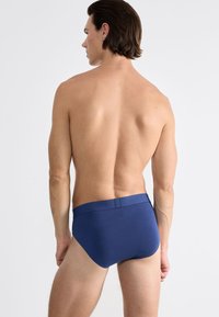 Mann mit schulterlangen Haaren, von hinten gesehen, in blauen Unterhosen vor einem schlichten hellen Hintergrund.