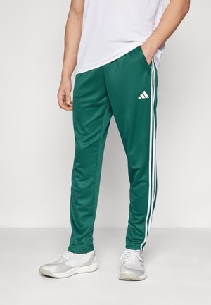 Grønne træningsbukser lavet af glat materiale med hvide sidelister og et Adidas-logo på venstre lår, udstyret med sidelommer.