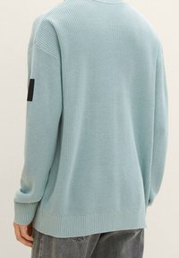 Osoba nosící světle modrý pletený svetr s černou nášivkou na levém rukávu a tmavě šedé džíny, zobrazená zezadu.