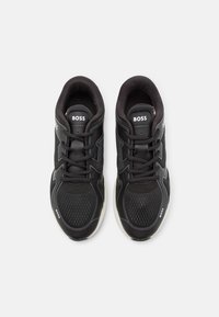 Chaussures de sport noires avec une tige en maille, une semelle texturée et un col rembourré. Les caractéristiques incluent des lacets et un logo sur la languette et le côté.