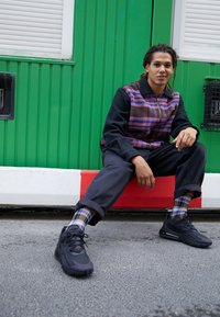 Jeune personne aux cheveux tressés assise sur un trottoir, portant une veste à carreaux, un pantalon sombre retroussé, des chaussettes à motifs et des baskets noires.