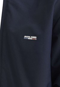 Veste navy avec un petit logo argenté et des accents bleu et rouge. Texture lisse et design minimaliste. Idéale pour une tenue décontractée.