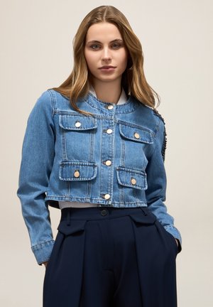 Veste en jean bleu cropped avec quatre poches avant, boutons dorés et col rond. Présente des détails texturés sur une épaule.