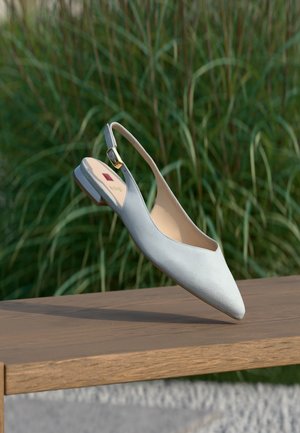 Zapato plano slingback azul claro con hebilla dorada, punta puntiaguda, descansando sobre una superficie de madera con fondo de hierba verde alta.