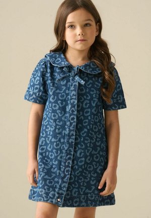 Junges Mädchen mit langen braunen Haaren, das ein blaues Kleid mit Leopardenmuster, kurzen Ärmeln und einem gebundenen Kragen trägt, steht vor einem schlichten Hintergrund.