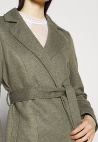 Zelený kabát s opaskem vyrobený z texturované látky, s širokými klopami, částečně přeložený přes lehkou košili s puntíky.