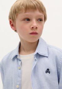 Lichtblauw gestreept overhemd met knoopsluiting, gedragen over een wit T-shirt, met een kleine geborduurde doodskop en gekruiste benen op de borst.