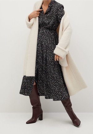 Abito nero a fiori con bottoni frontali, indossato sotto un oversize cardigan lavorato a maglia crema. Stivali bordeaux al ginocchio completano il look.