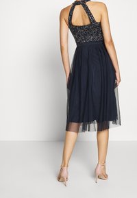 Mekko, jossa on kiiltävä halterneck-pääntie, polven pituinen tyllihelma ja istuva vyötärö. Ominaisuuksiin kuuluu avokanta selässä ja vaaleanpunaiset nilkkaketjupohjaiset korkokengät.