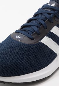 Marineblaue Adidas Sneakers mit weißen Streifen, marinefarbenen Schnürsenkeln, gestricktem Obermaterial und weißen Zwischensohlen, im schrägen Nahaufnahme der Zehen- und Schnürbereich.