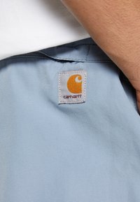 Gros plan sur une poche de vêtement bleu clair avec un patch logo Carhartt blanc et orange. Le bras d'une personne est partiellement visible.