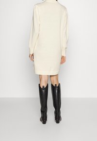 Femme portant une robe pull crème à manches longues et des bottes en cuir noires jusqu'au genou, se tenant devant un fond léger uni.