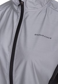 Lysegrå vindjakke med sort lynlås og detaljer. Har en glat tekstur og "ENDURANCE" logo trykt på forsiden.