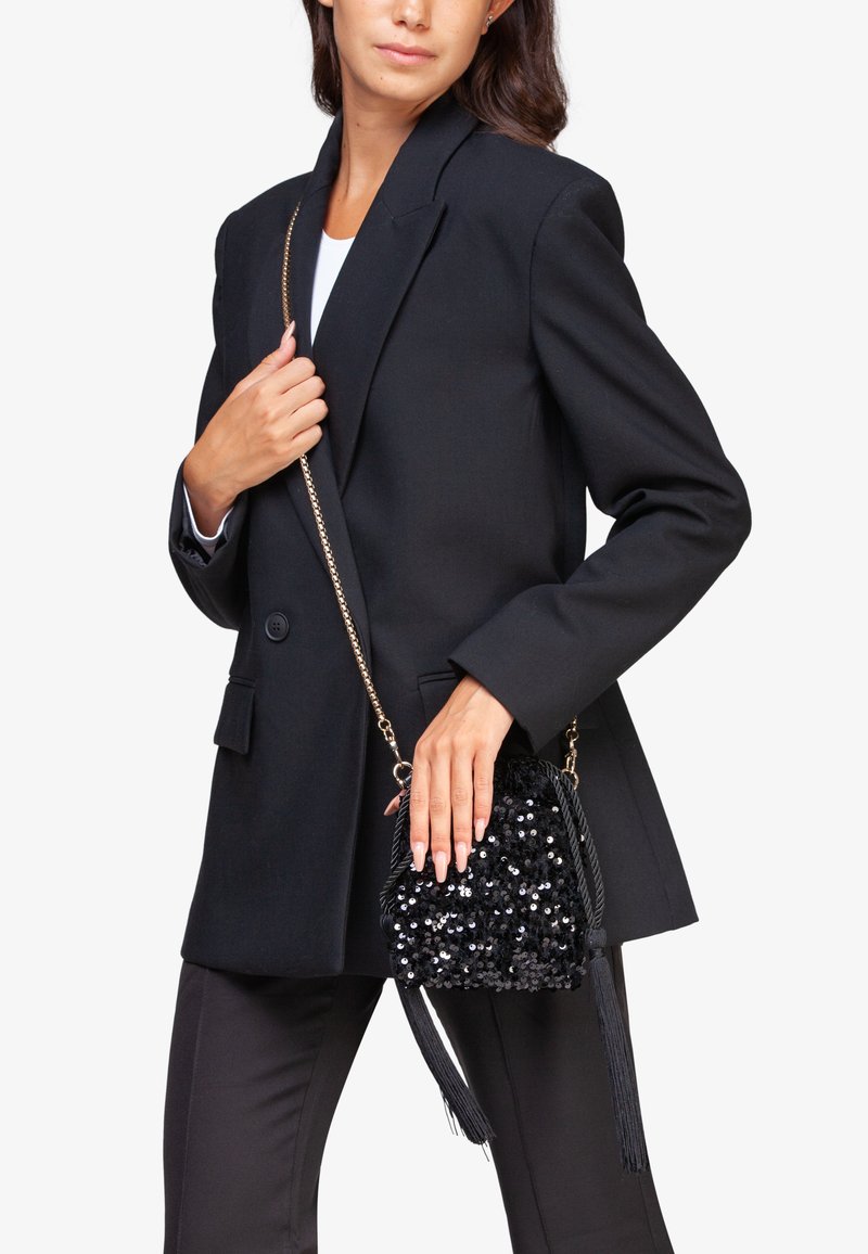 Bolso cruzado negro con lentejuelas, cierre de cordón, borlas y una correa de cadena, sostenido por una persona que lleva un blazer negro.